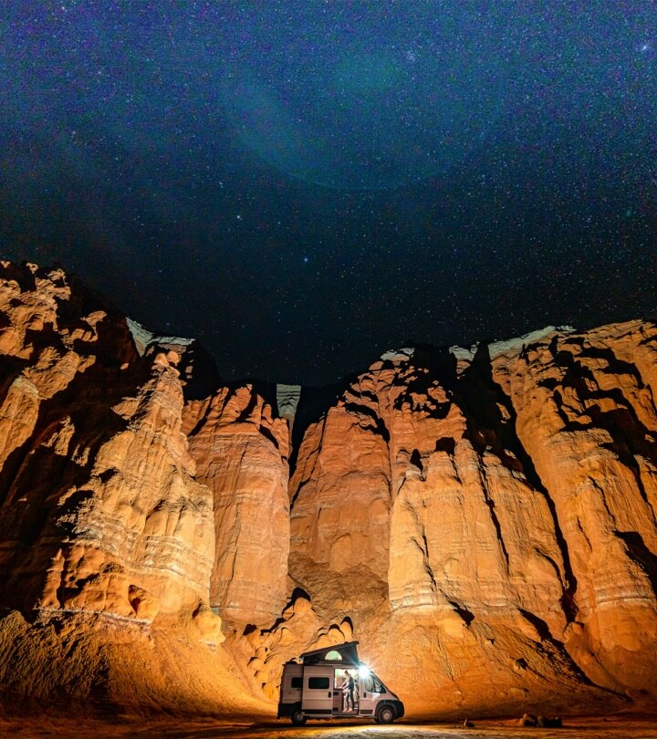 White van parked below a red rock wall emitting light which bounces off the wall with the starry sky shown above.