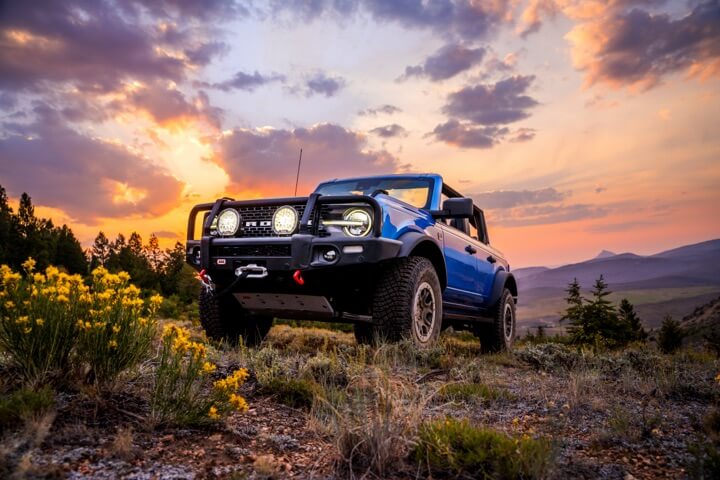 A Blue Ford Bronco truck with ARB accesory lights, front bumper, front hitch with a sunset in the background and yellow desert flowers in foreground.